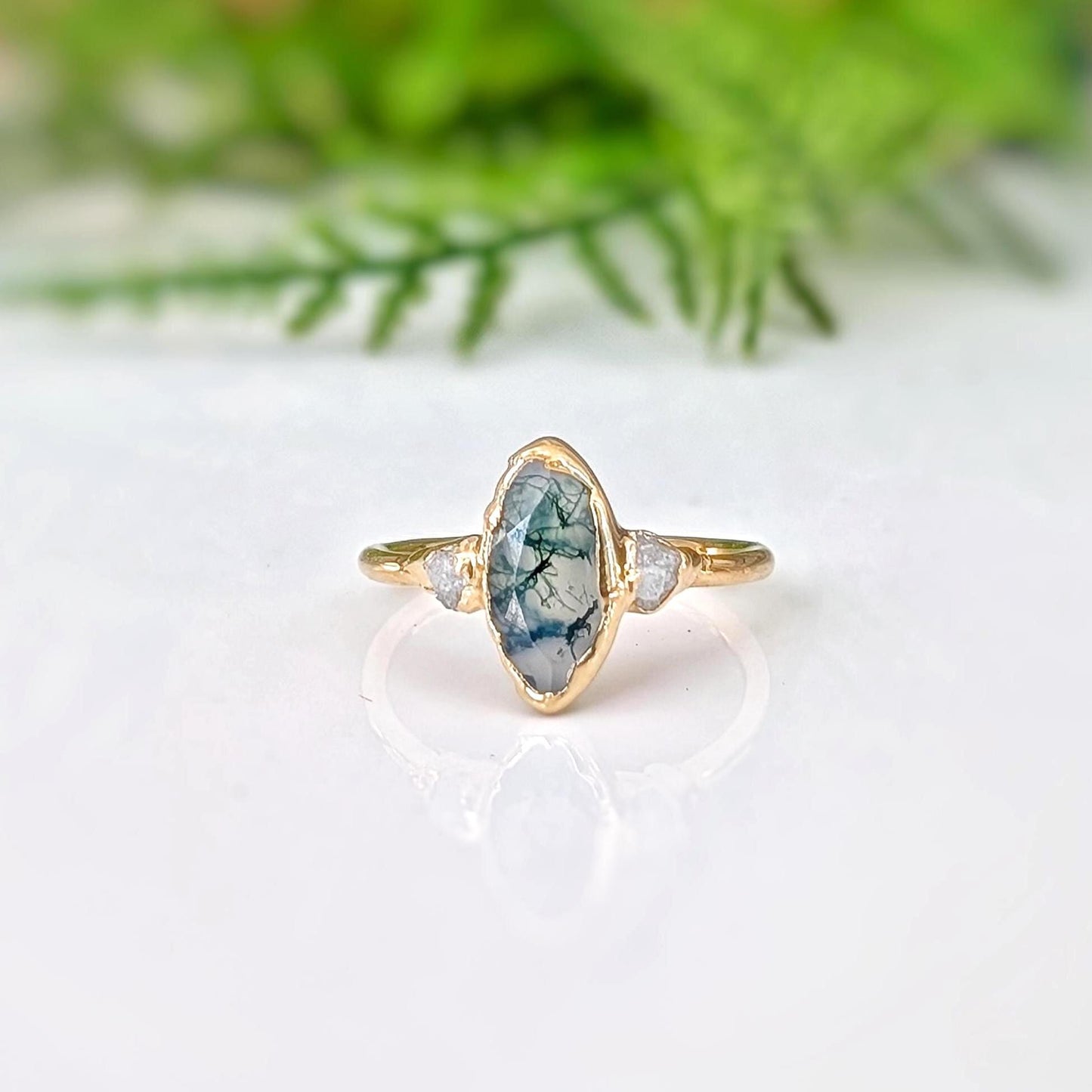 Gold ring with a marquise shape Moss Agate and raw diamond engagement ring in a light background with some greenery