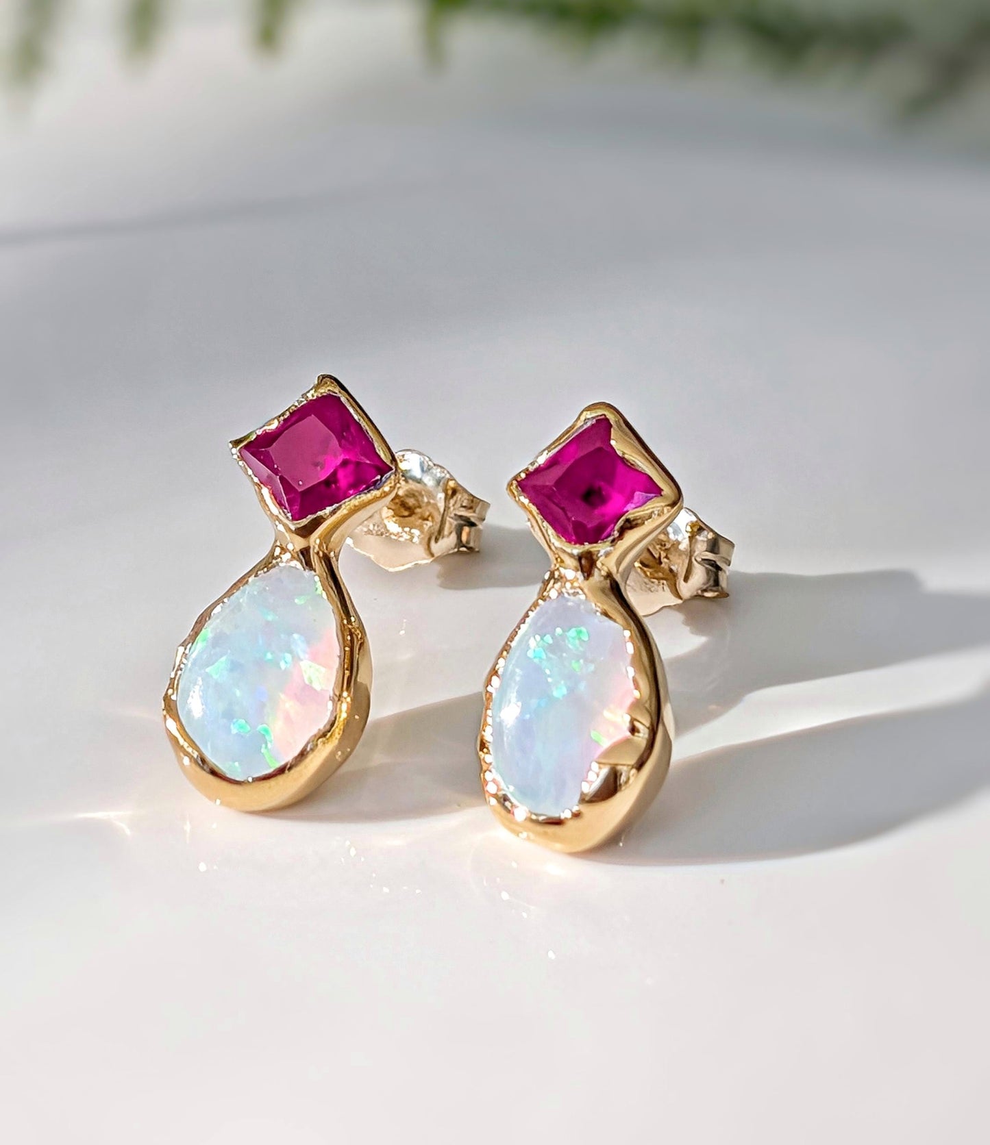Gold stud earrings with dark pink Ruby and white Opal gemstones on a light background