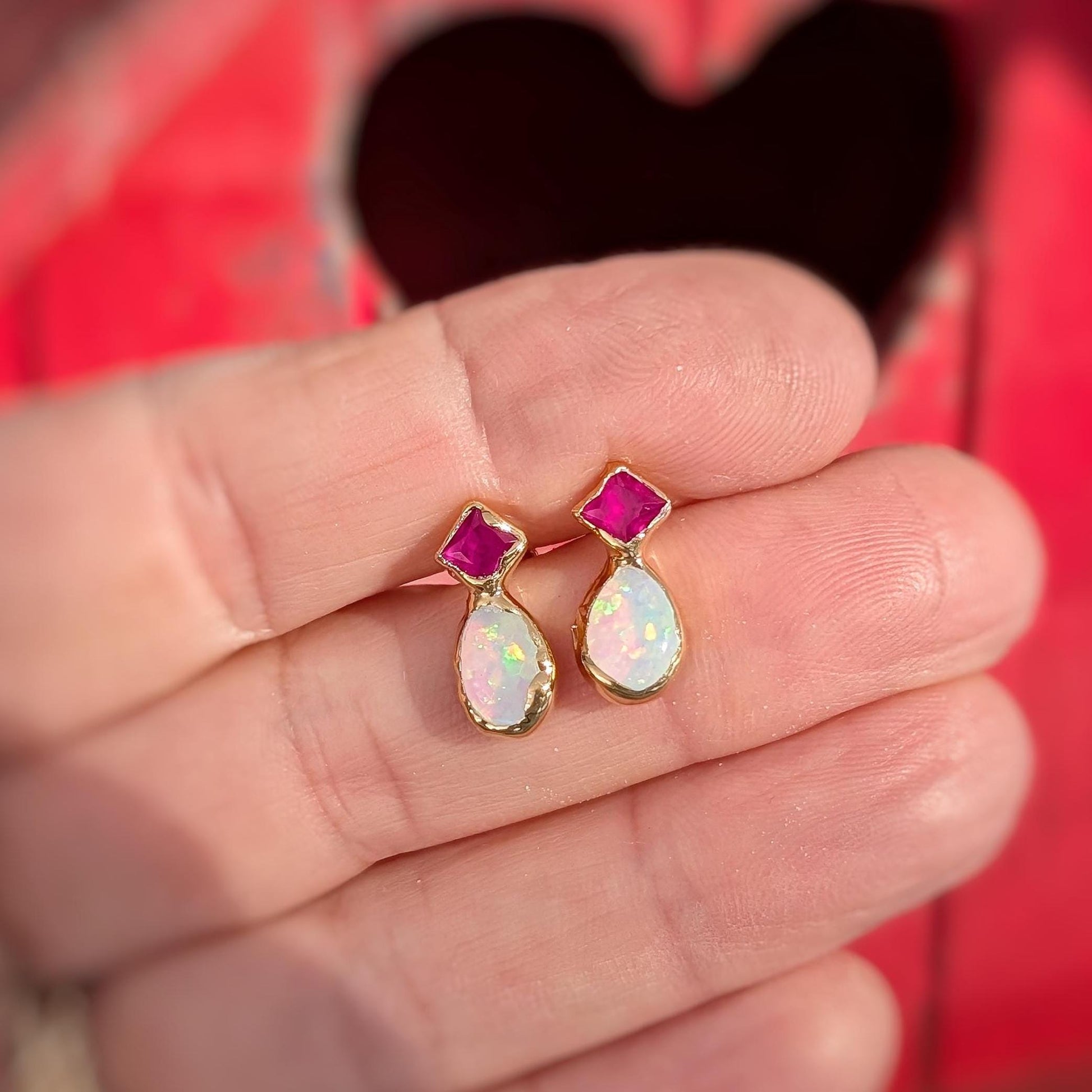 A hand holding a Pair of Gold stud earrings with dark pink Rubies and White Opals