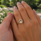 A gold ring with a central pearl and small surrounding clear stones, shown worn on a person's finger with hands in front of a blurred outdoor background.
