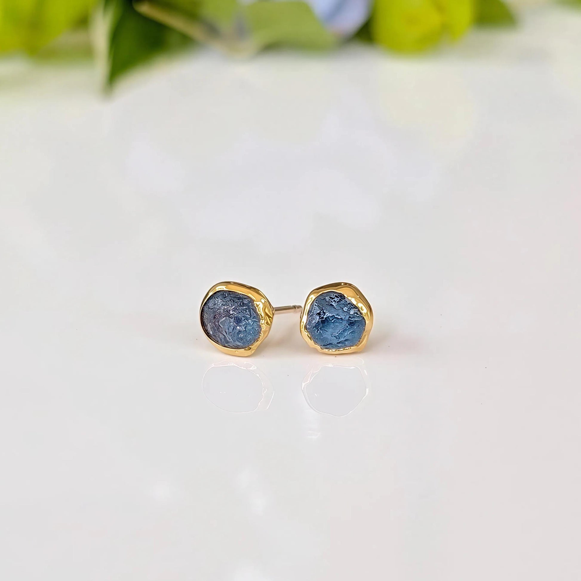 A pair of gold earrings with raw blue Montana Sapphires - sitting on a white surface, with green leaves in the background.