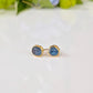A pair of gold earrings with raw blue Montana Sapphires - sitting on a white surface, with green leaves in the background.