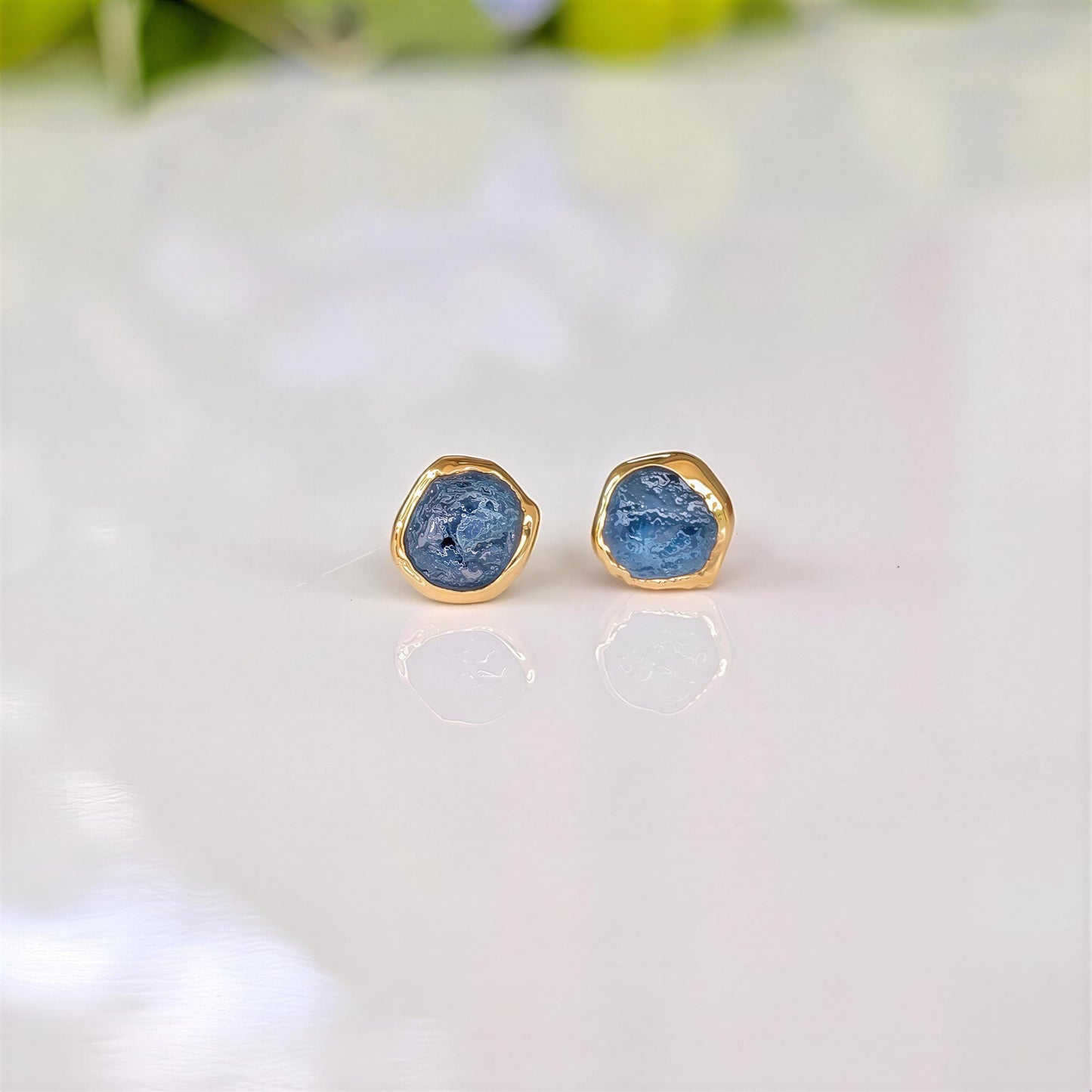 A pair of gold stud earrings with raw blue Montana Sapphires, placed on a white surface.