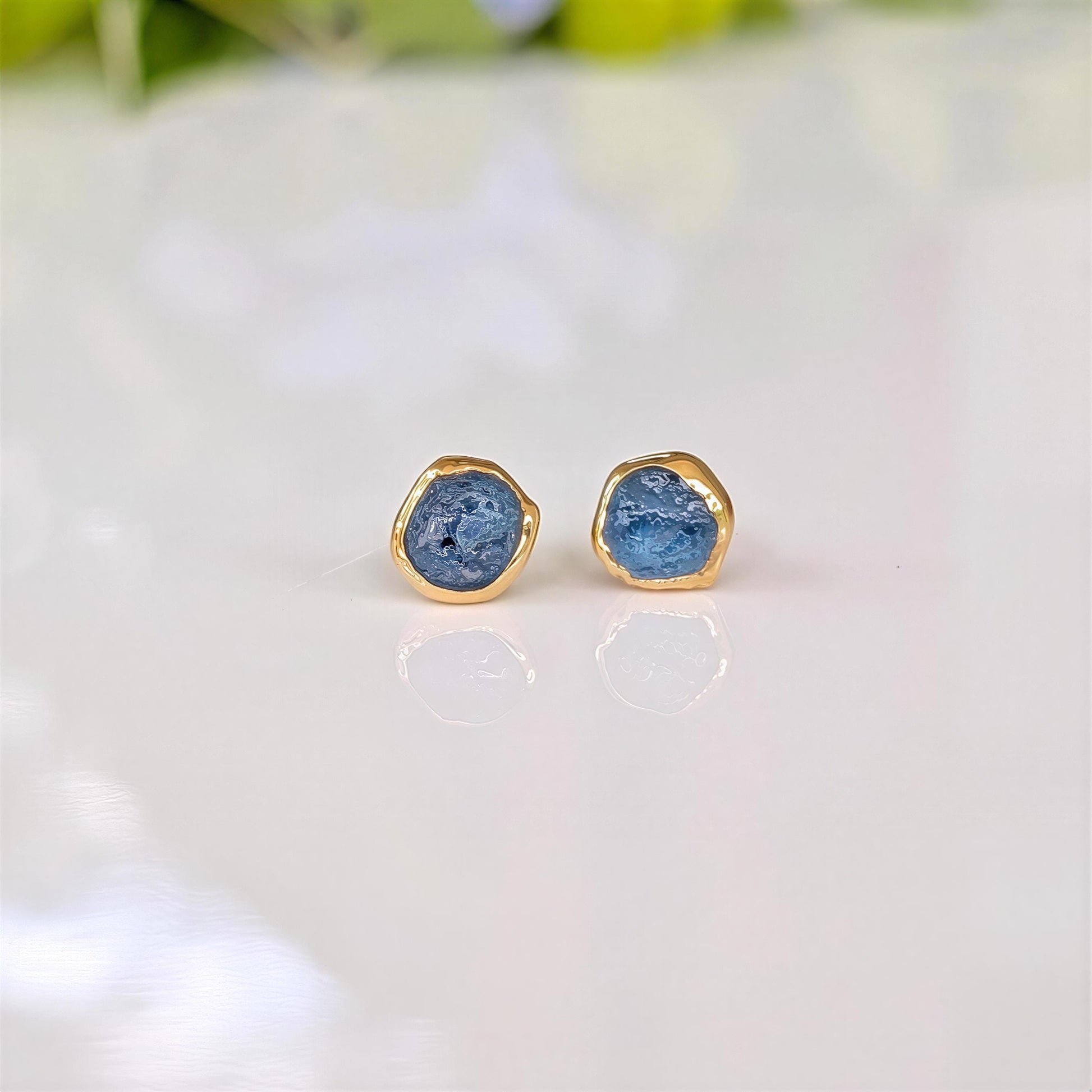 A pair of gold stud earrings with raw blue Montana Sapphires, placed on a white surface.