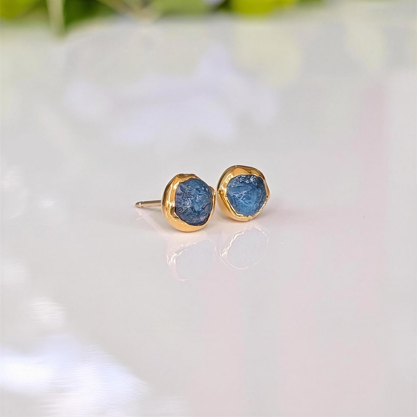 A pair of gold stud earrings with raw blue Montana Sapphires, placed on a white surface.