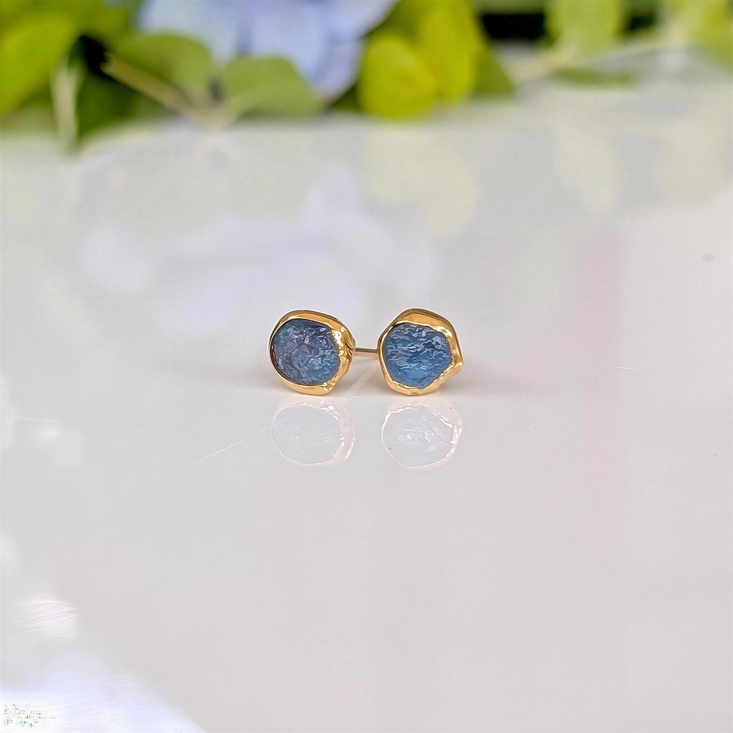 A pair of gold stud earrings with raw blue Montana Sapphires, placed on a white surface.