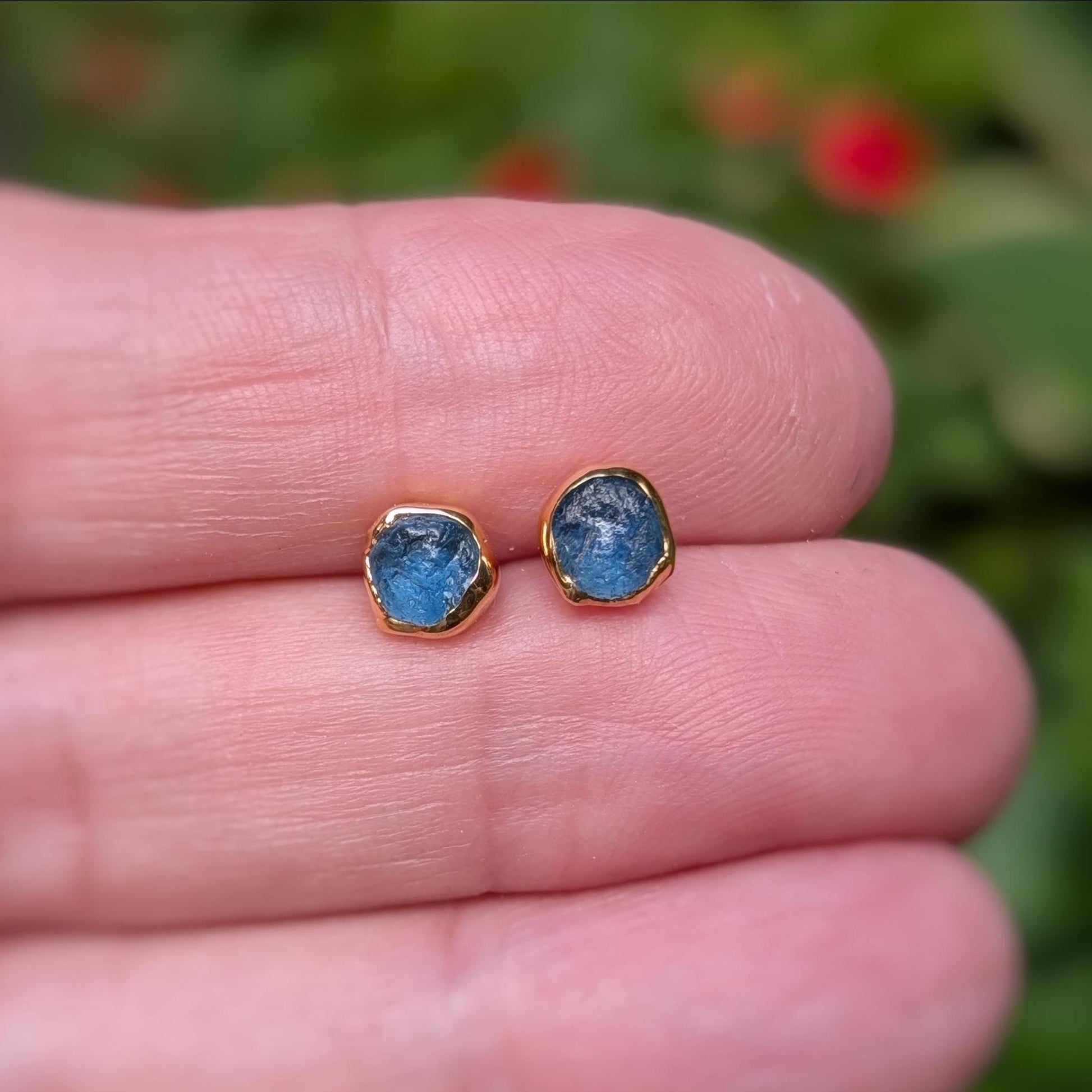A hand holding a pair of gold stud earrings with raw blue Montana Sapphires