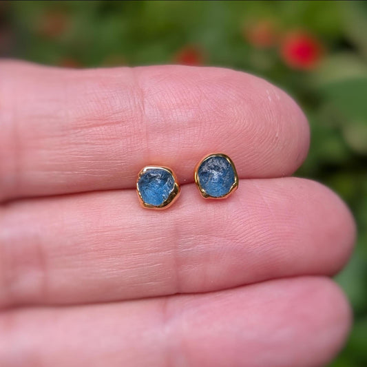 A hand holding a pair of small, raw Teal blue Montana Sapphire stud earrings.