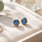A pair of Teal blue Montana Sapphire stud earrings with textured gold bezel settings, displayed on a beige textured jewelry tray with a gold ring and eucalyptus leaves nearby.