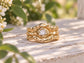 A gold-textured wedding ring set with a large central Moissanite sits on a wooden surface, surrounded by white flowers and leaves.