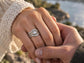 A close-up image of a woman's hand wearing a Silver Textured wedding ring set featuring a large round Moissanite, with another man's hand gently holding the woman's hand.