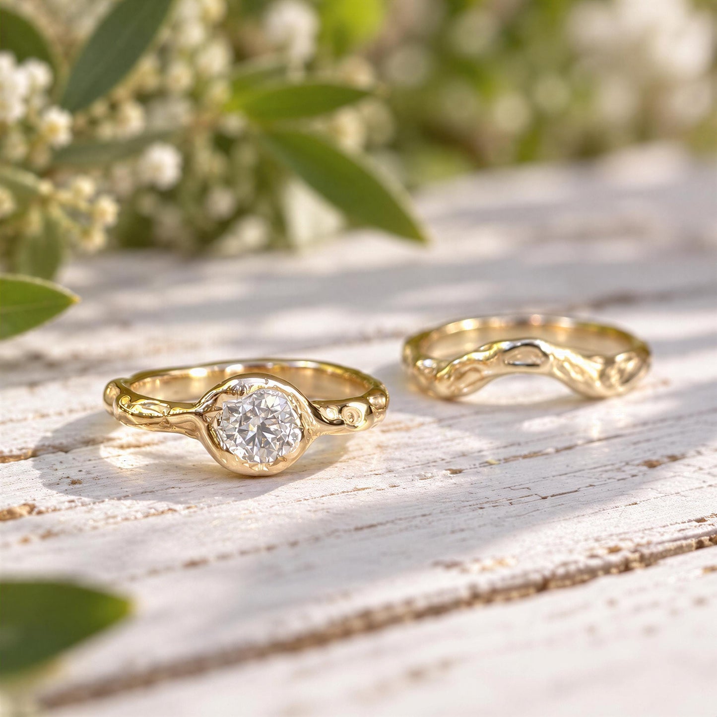 Two gold rings with a large central Moissanite are placed on a wooden surface, with a blurred background of greenery.
