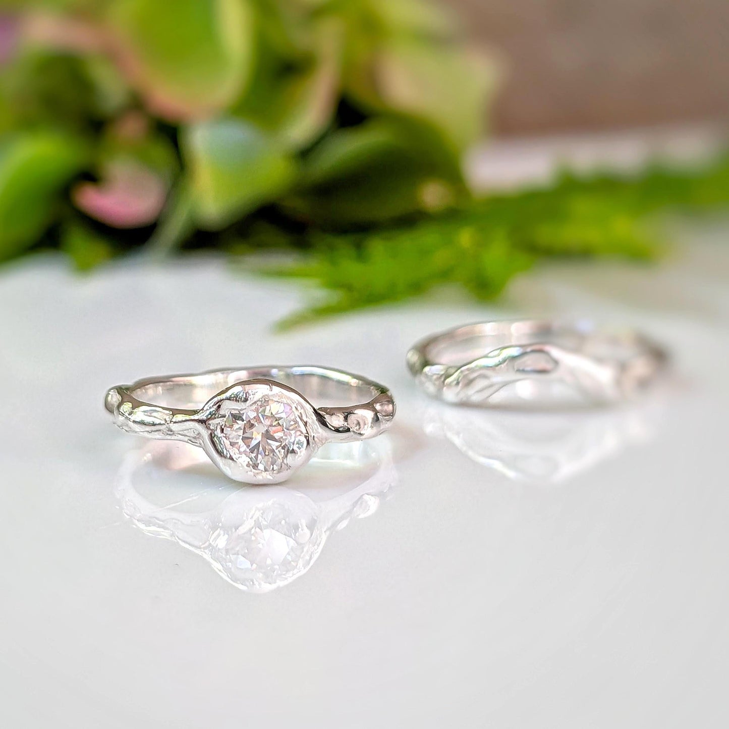 Two silver rings with a large central Moissanite are displayed on a white surface, with a blurred green plant in the background.