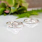 Two silver rings with a large central Moissanite are displayed on a white surface, with a blurred green plant in the background.