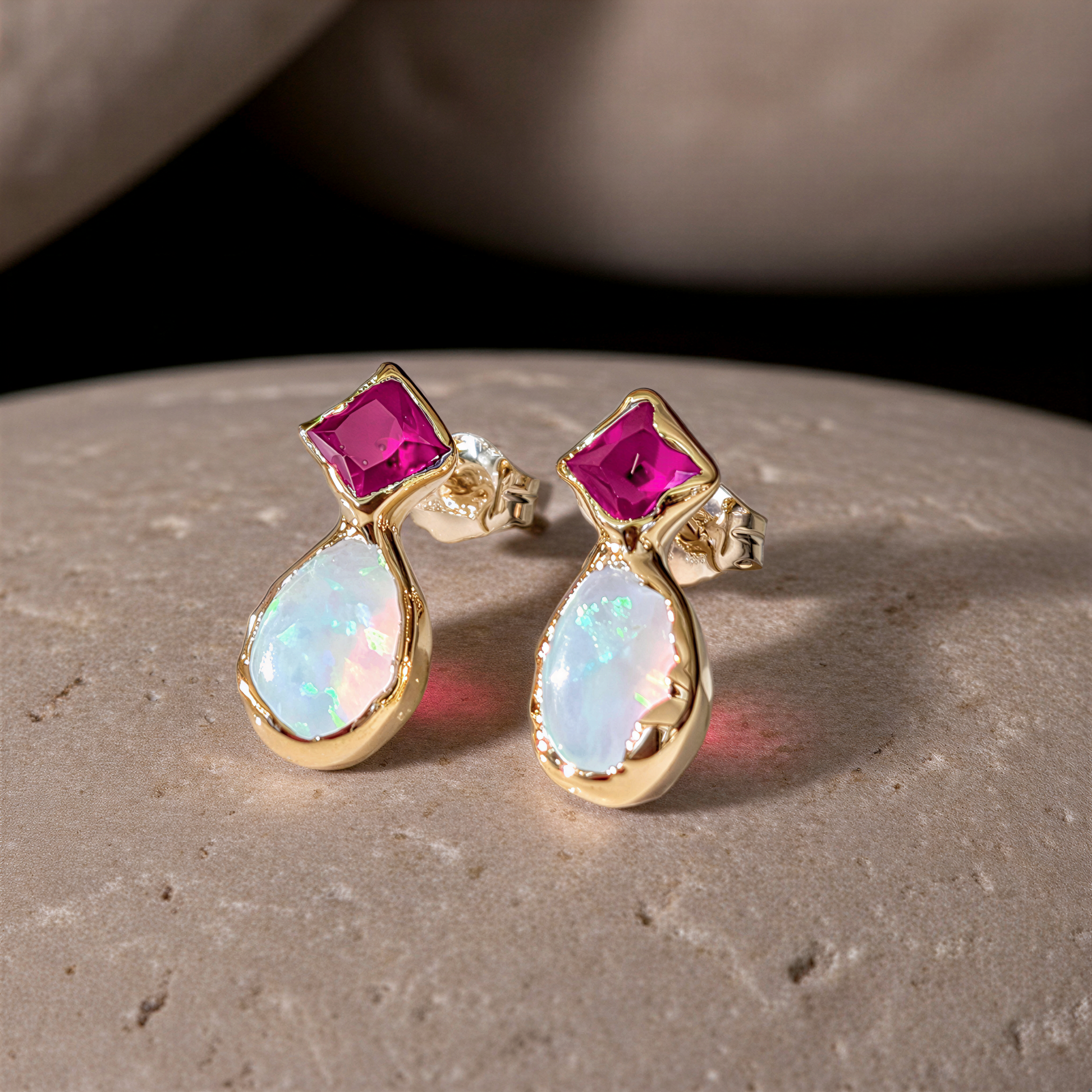 Gold earrings with a dark pink Ruby and opal gemstones on a stone surface