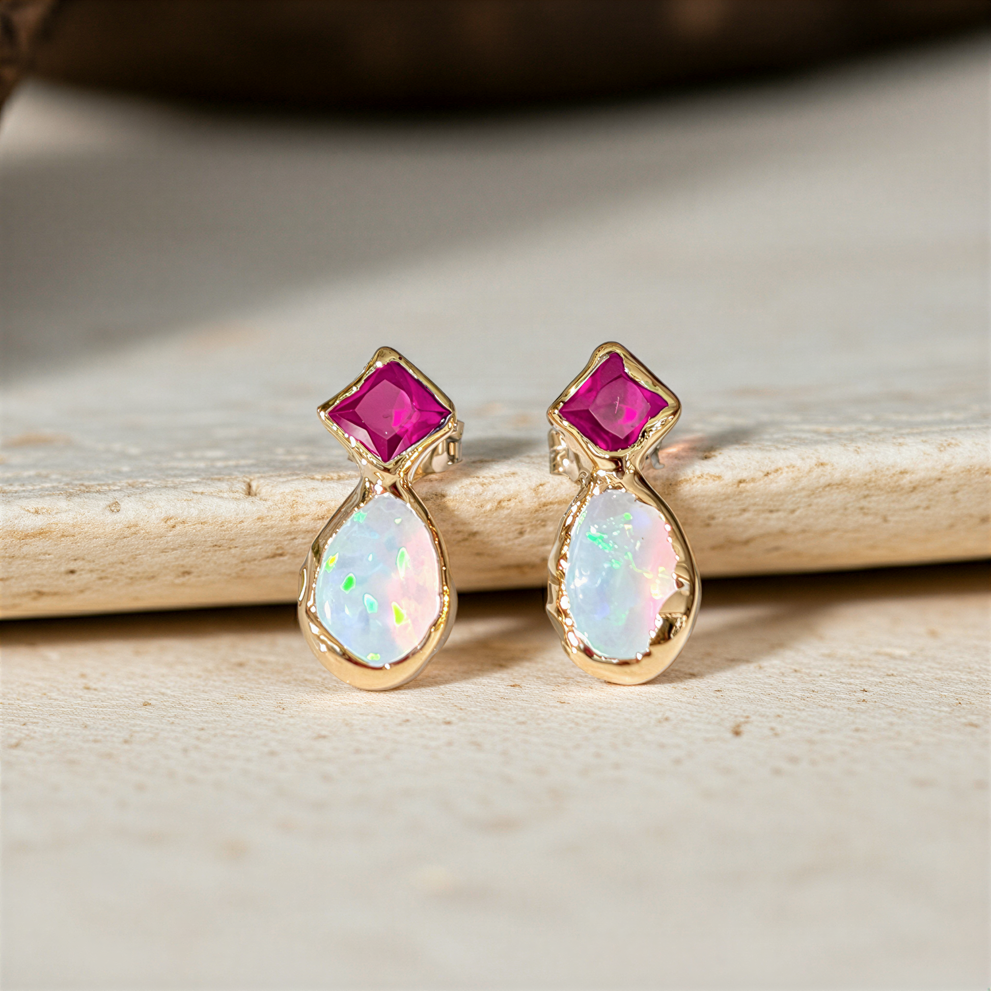 Gold stud earrings with dark pink Ruby and Opal gemstones on a wooden surface