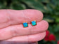 A hand holding a pair of square shape flashy blue & green Opal stud earrings in Gold organic setting