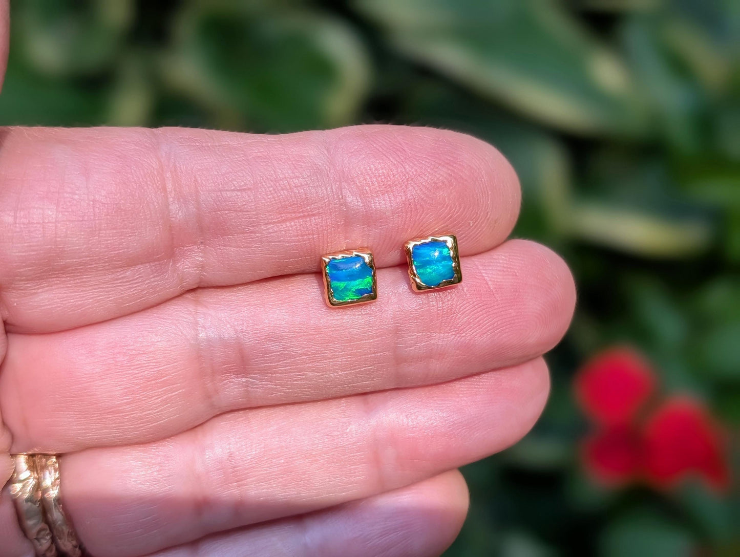 A hand holding a pair of square shape flashy blue & green Opal stud earrings in Gold organic setting