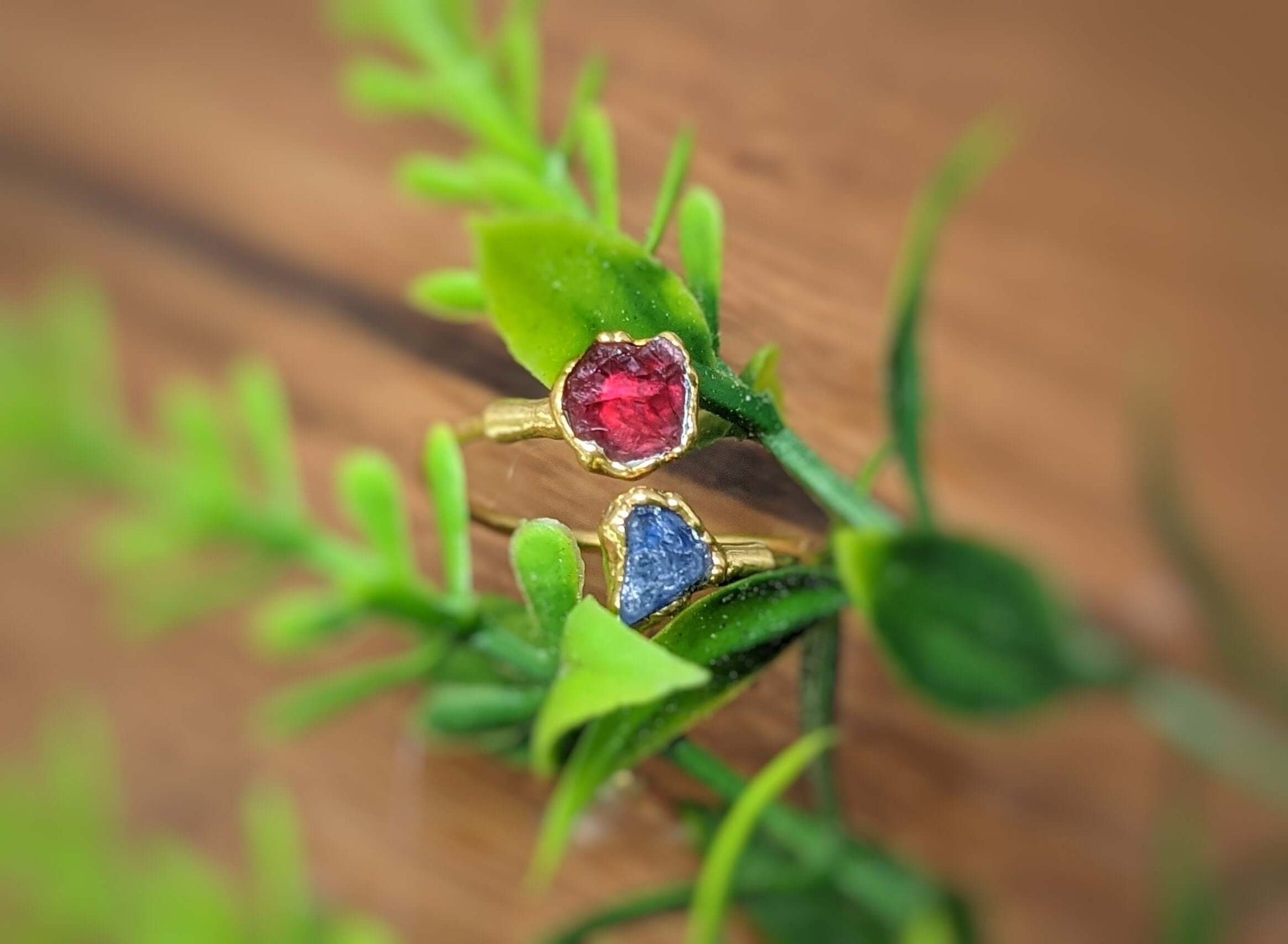 Toi et Moi raw gemstone rings in gold featuring a red and blue heart-shaped gemstone, set against a green backdrop.
