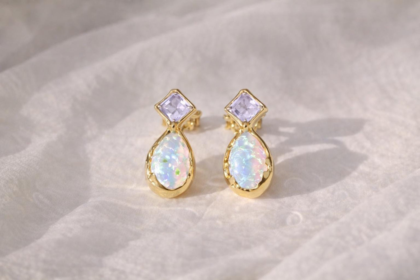 Pair of gold stud earrings with Amethysts and opal gemstones on a textured beige surface