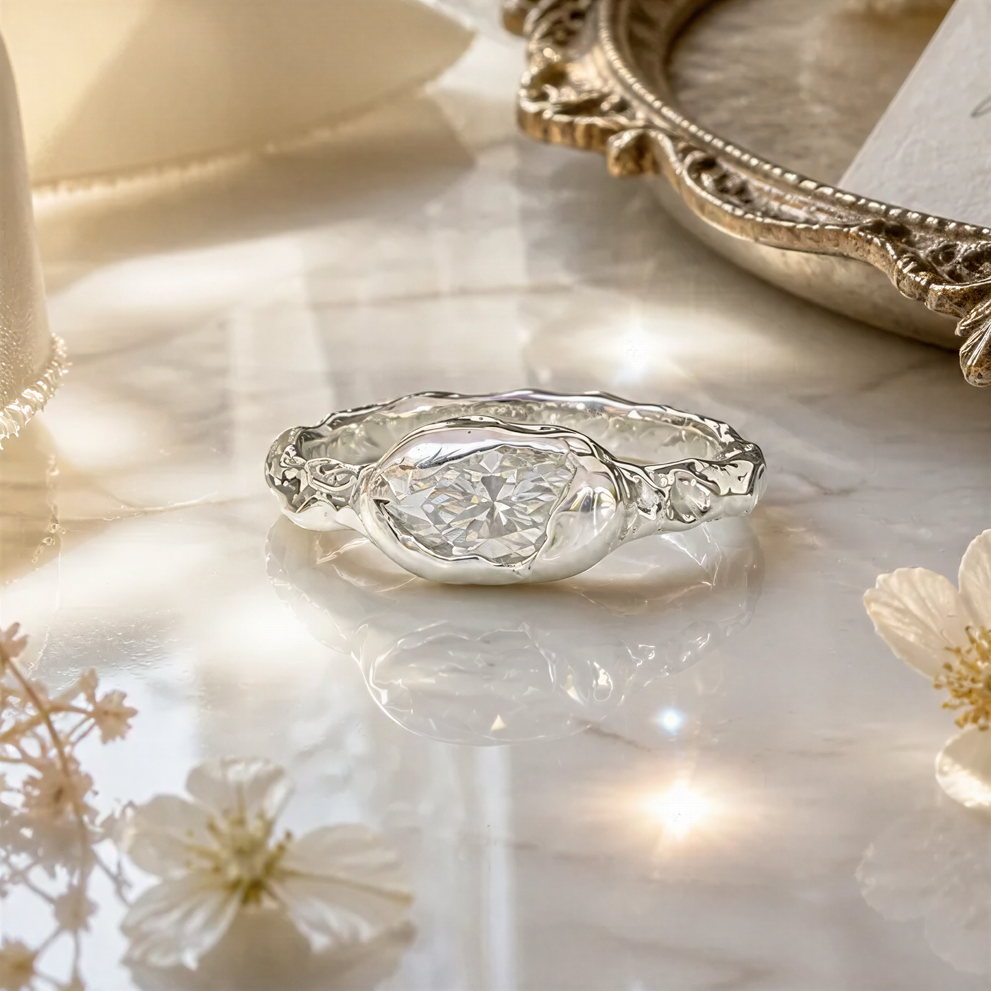 A silver ring with a large diamond set in the center, surrounded by a twisted band, sits on a marble surface with a silver tray and white flowers in the background.
