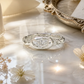 A silver ring with a large diamond set in the center, surrounded by a twisted band, sits on a marble surface with a silver tray and white flowers in the background.