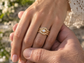 A close-up image of a person's hand wearing a gold ring with a diamond, with another person's hand gently holding the ring.