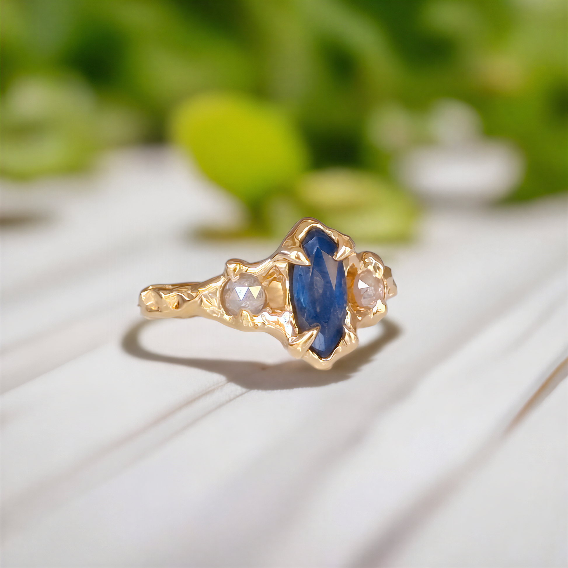 A gold ring featuring a marquise-cut blue sapphire center stone and two small round clear stones, resting on a white wooden surface with a soft-focus green background.