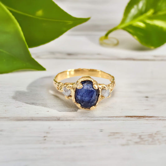 Blue sapphire and diamond engagement ring in solid gold with textured band, surrounded by green foliage.