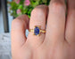 Hand wearing a blue sapphire ring in gold, featuring an emerald cut stone and intricate band design.