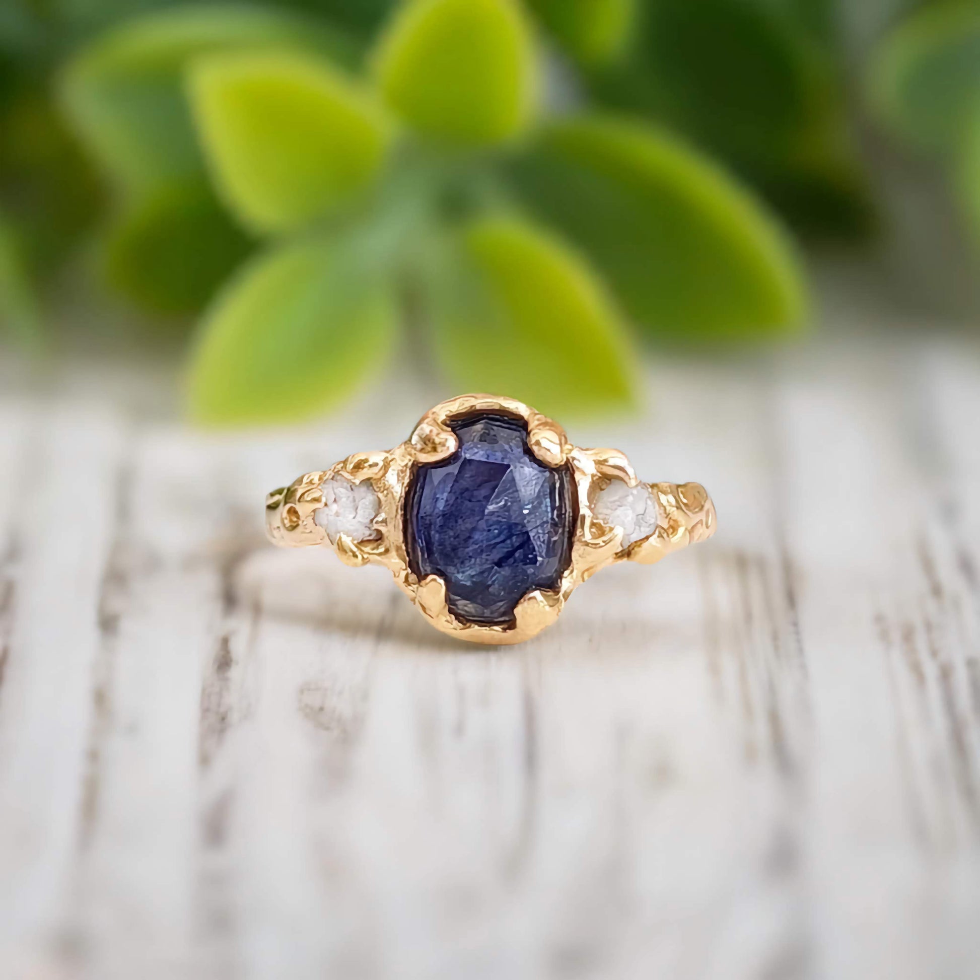 Blue Sapphire and raw diamond engagement ring with gold setting on wooden surface.