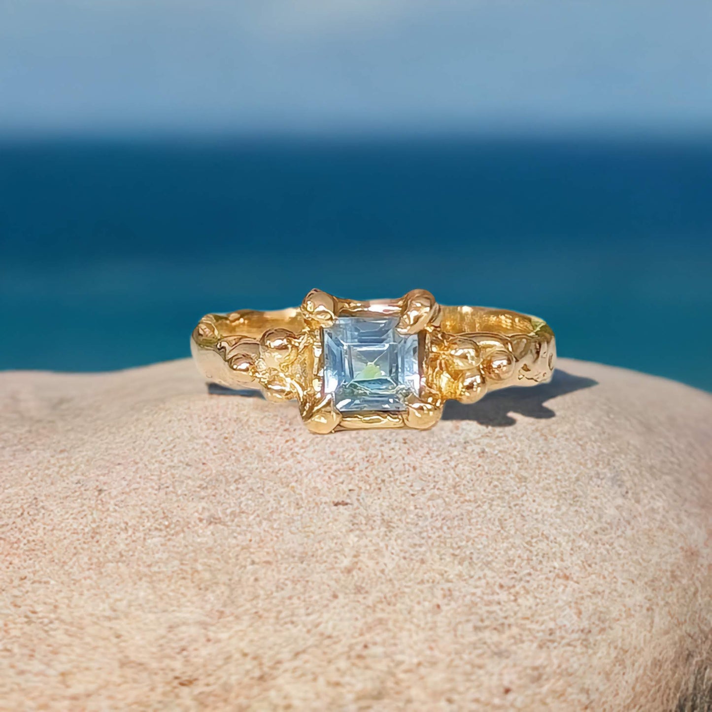 Solid 14k Gold engagement ring featuring a London Blue Topaz gemstone on a textured band, set against a beach background.