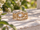 A gold ring with a large central diamond set in a unique, organic design sits on a wooden surface, surrounded by green foliage.
