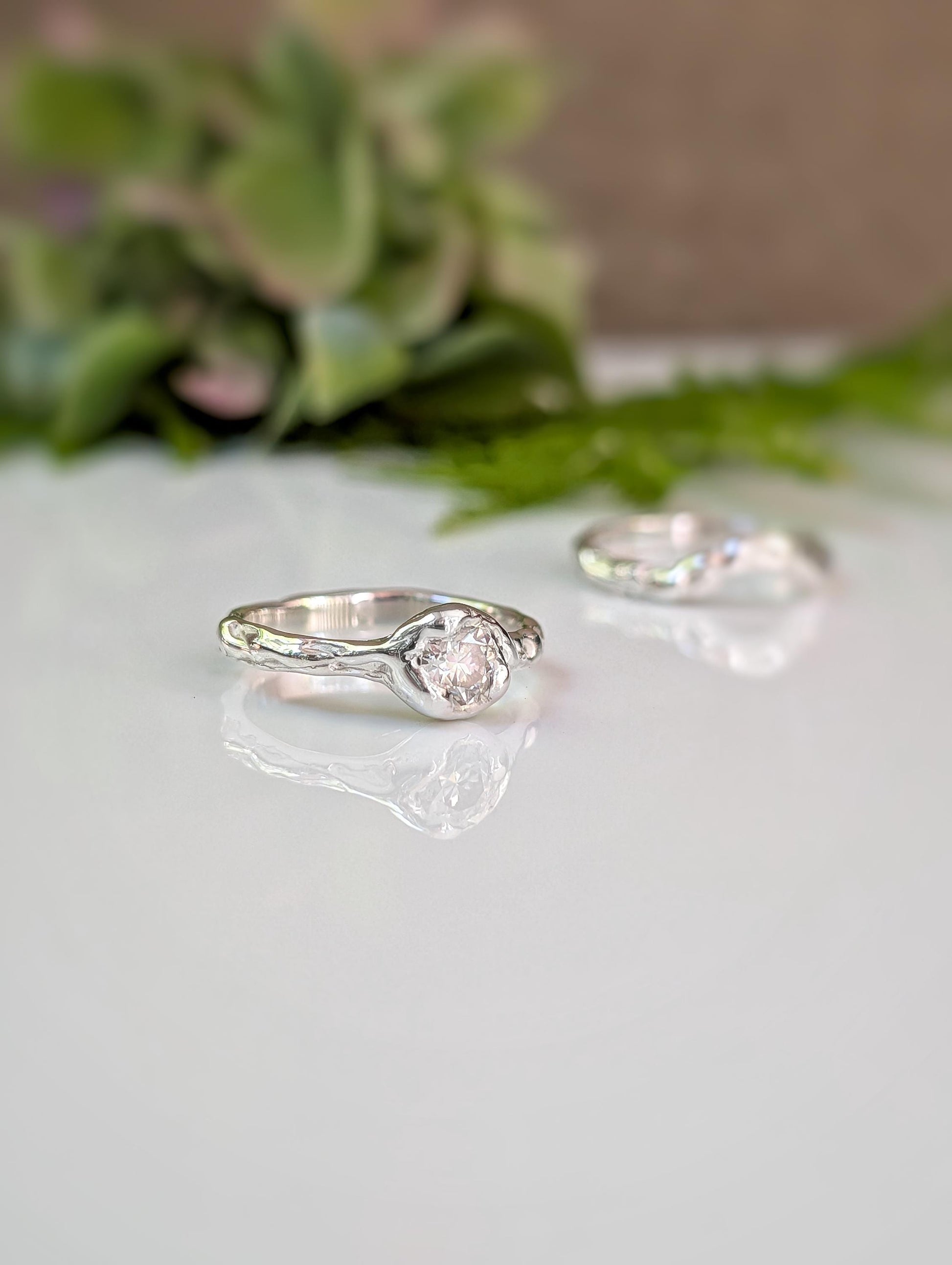 Two silver rings with a central stone are displayed on a white surface, with a blurred green plant in the background.