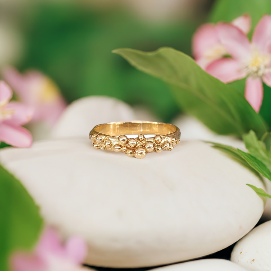 Solid gold Dew Drop Molten beaded ring on smooth stones surrounded by pink flowers, showcasing its unique organic design.