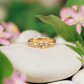 Solid gold Dew Drop Molten beaded ring on smooth stones surrounded by pink flowers, showcasing its unique organic design.