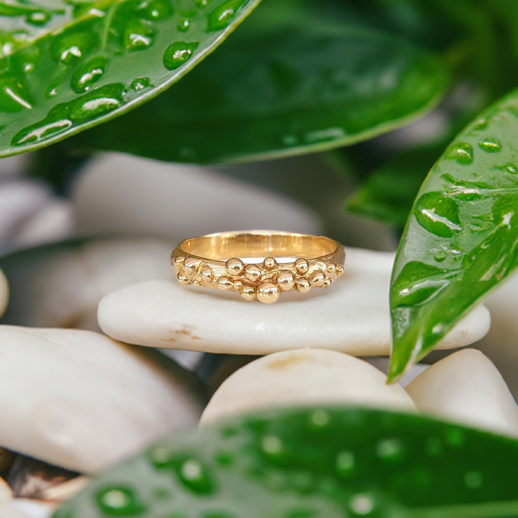 Solid Gold Dew Drop beaded ring resting on smooth stones amidst lush green leaves with water droplets.