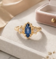 A gold ring with a blue gemstone, placed on a white surface, with a pink velvet box and some flowers in the background.