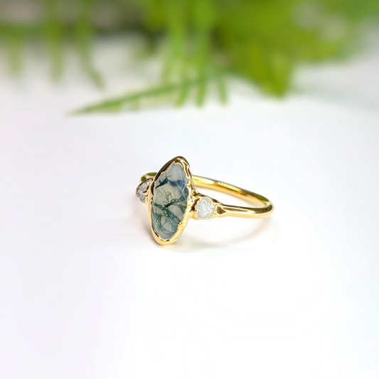 Side view of a Gold Moss Agate and raw diamond engagement ring in a light background with some greenery