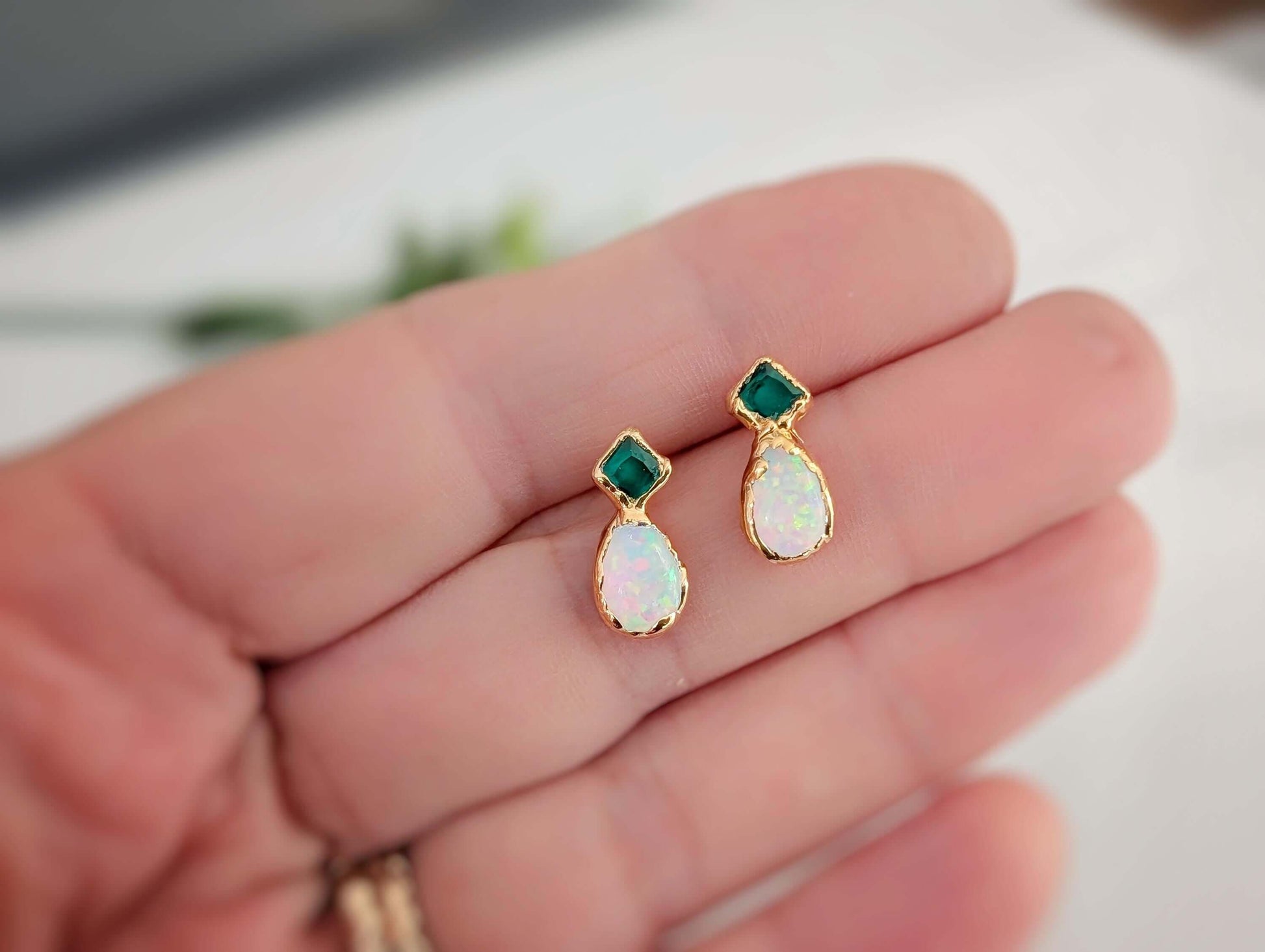 A hand holding a pair of Gold earrings with green Emerald and Opal