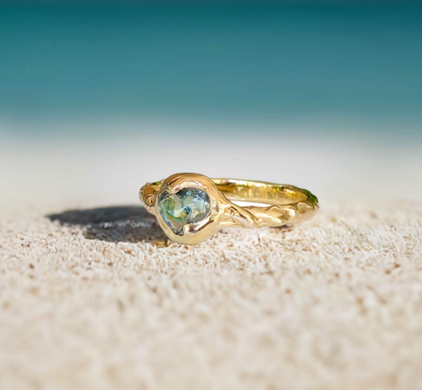 Australian Sapphire ring in solid 14k gold on sandy beach with ocean background, showcasing teal gemstone.