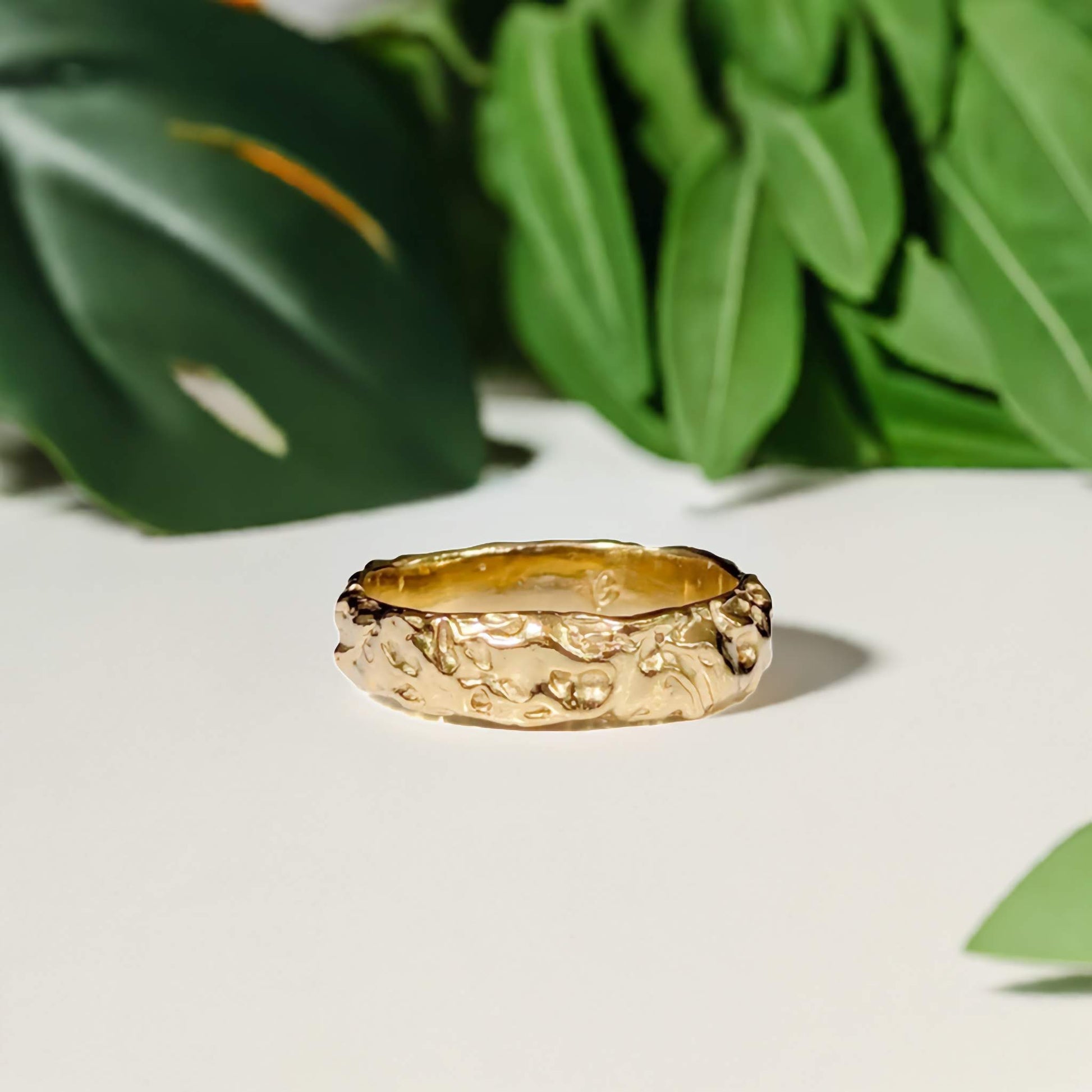Handcrafted molten gold wedding ring featuring a rustic textured design, surrounded by green leaves.