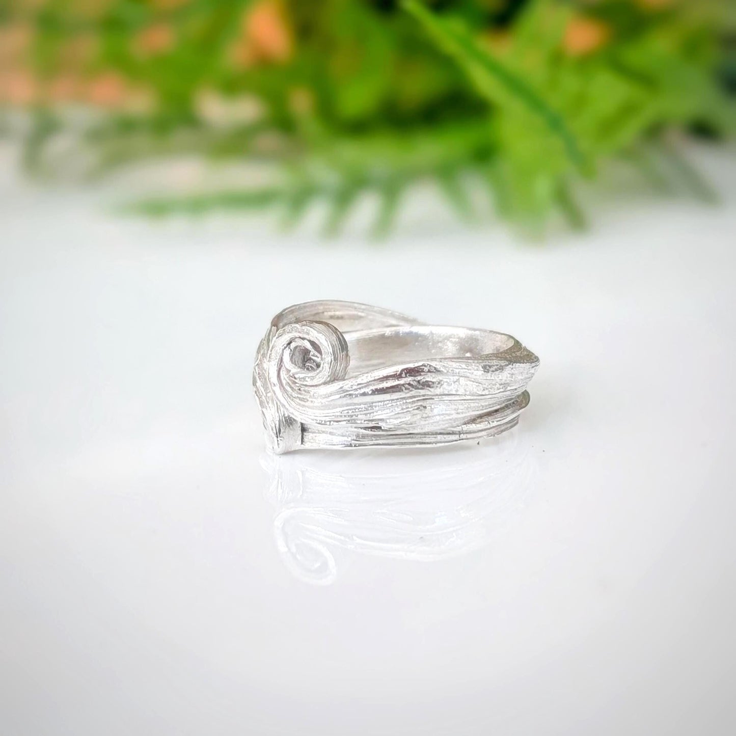 A silver ring with a textured, organic swirl design, resting on a white reflective surface with a soft-focus green background.