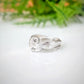 A silver ring with a textured, organic swirl design, resting on a white reflective surface with a soft-focus green background.