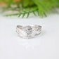 A silver ring with a textured, organic swirl design, resting on a white reflective surface with a soft-focus green background.