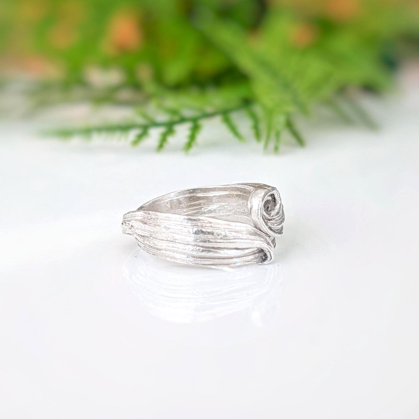 A silver ring with a textured, organic swirl design, resting on a white reflective surface with a soft-focus green background.