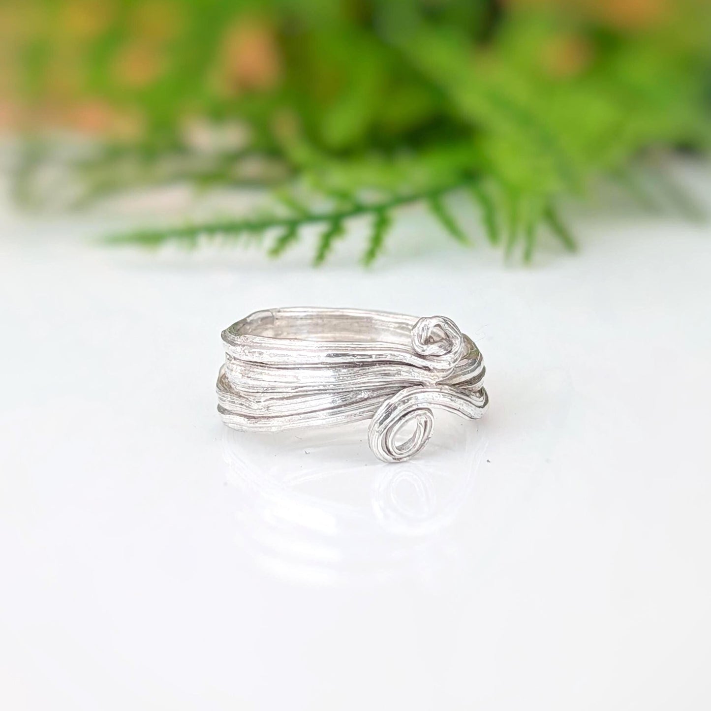 A silver ring with a textured, organic swirl design, resting on a white reflective surface with a soft-focus green background.