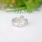 A silver ring with a textured, organic swirl design, resting on a white reflective surface with a soft-focus green background.
