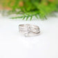 A silver ring with a textured, organic swirl design, resting on a white reflective surface with a soft-focus green background.