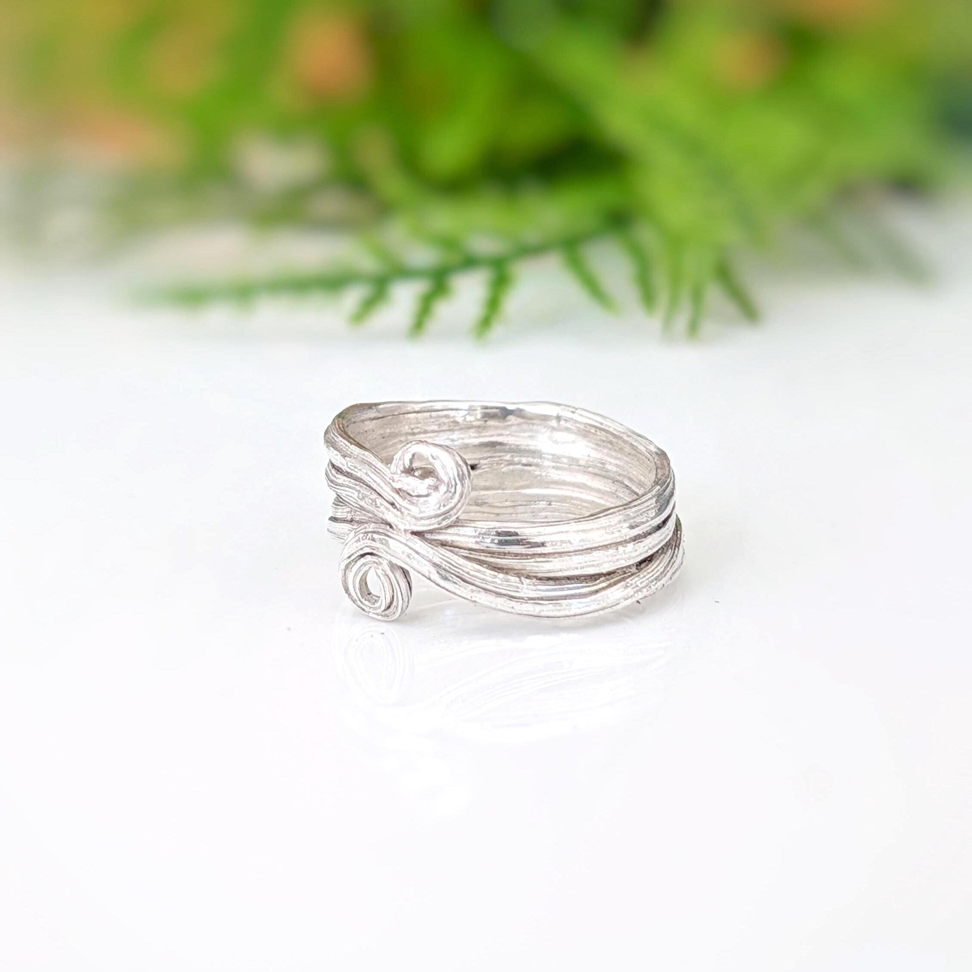 A silver ring with a textured, organic swirl design, resting on a white reflective surface with a soft-focus green background.